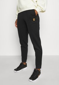Černé tapered joggery z měkké tkaniny, s boční kapsou a malým barevným logem na přední straně. Noseny s černými teniskami.