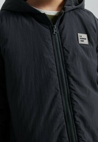 Must kapuutsiga jakk tekstuurse viimistlusega, millel on ees tõmblukk ja rinnal logomärk. Pehme sisemusega nähtav vooder.