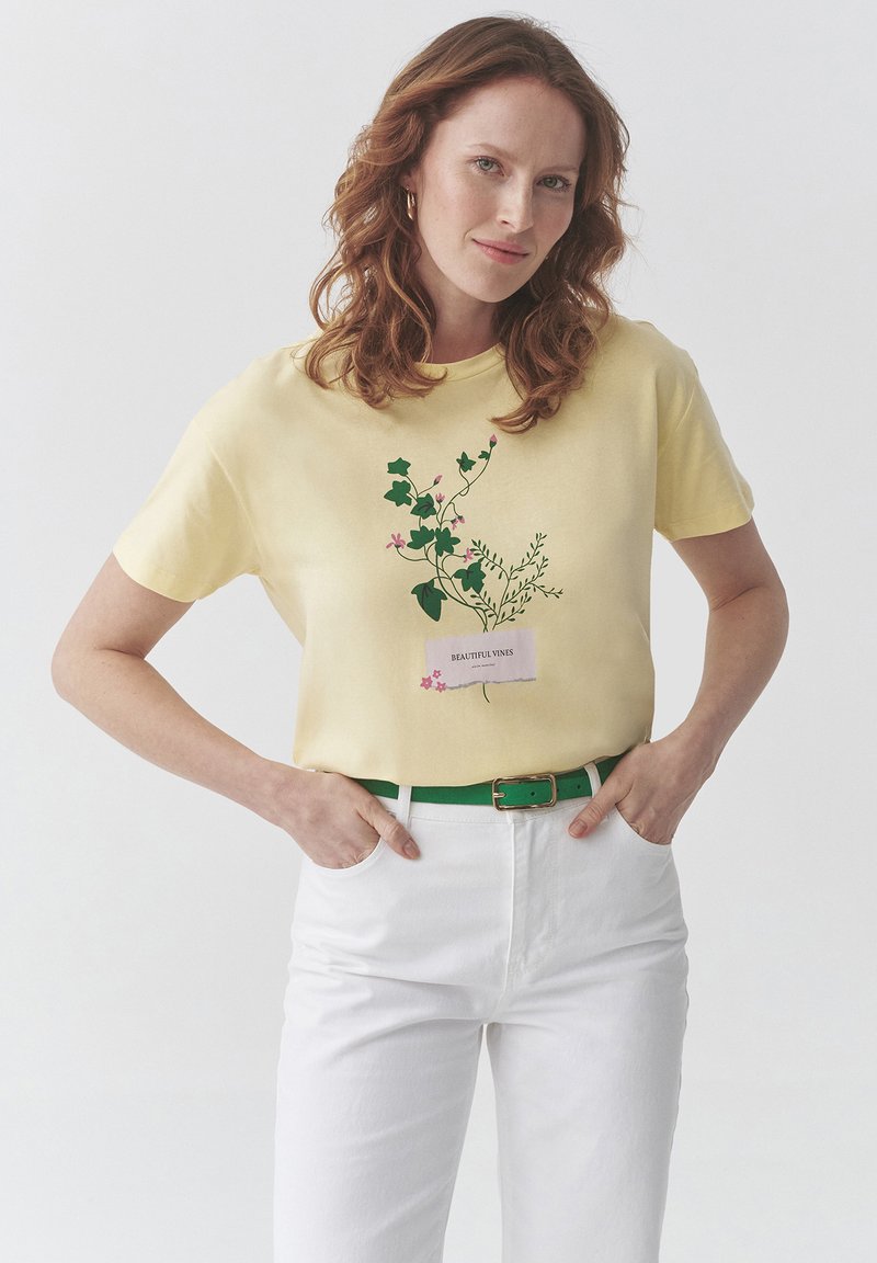 Kvinde med rødt hår iført en gul skjorte med grønne vinranker og rosa blomsterprint, hvide bukser og et grønt bælte, hænderne i lommerne.