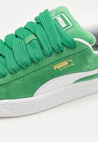 Grønn semsket sneaker med tykke, teksturerte snørebånd, en hvit gummisåle og gullfargede merkevarer. Rund form med kontrasterende detaljer.