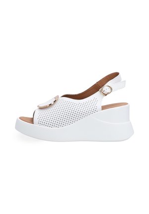 Hvid kile-sandal med perforeret overdelsmateriale, åben tå, slingback-rem med gyldent spænde og dekorativt gyldent cirkulært accent på toppen.