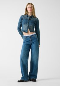 Τζάκετ cropped από τζιν σε βαθύ μπλε με κούμπωμα με κουμπιά και κάθετες ραφές, συνδυασμένο με τζιν παντελόνι φαρδιάς γραμμής σε μπλε χρώμα με λευκή μέση με λογότυπο.