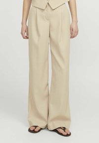 Beige wijde broek van een zachte stof, met een hoge taille, plooien en een stijlvolle snit. Draagt met bruine teenslippers.