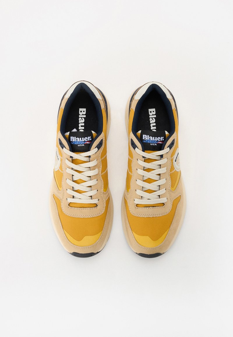 Gelbe und beigefarbene Sneaker mit strukturiertem Obermaterial, runder Zehe und weißen Schnürsenkeln; ausgestattet mit einer kontrastierenden schwarzen Sohle und einem gebrandeten Innenraum.