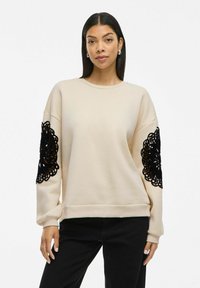 Beige sweatshirt with black lace detailing on sleeves, featuring a relaxed fit and crew neckline, made from soft fabric.