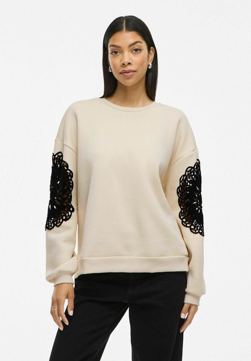 Beige sweatshirt with black lace detailing on sleeves, featuring a relaxed fit and crew neckline, made from soft fabric.