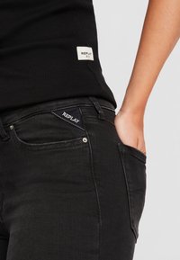 Jeans de denim negro con una silueta ajustada, herrajes plateados y un logo bordado "REPLAY" en el bolsillo. Top negro acanalado en la cintura.