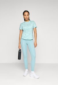 Lys blå athletic t-skjorte med et subtilt mønster, kombinert med matchende leggings. Modellen holder en svart vannflaske og har på seg hvite joggesko.