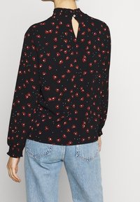 Blusa nera con piccolo motivo floreale rosso, caratterizzata da un colletto alto con dettaglio di volant e apertura a chiave sul retro. Maniche lunghe con arricciatura ai polsi.