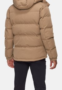 Hnědá péřová bunda s kapucí, se štepovaným designem a elastickými manžetami, oblečená přes tmavé kalhoty. Název značky je vyšité na zadní straně.