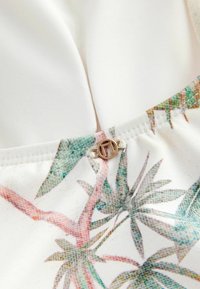 Vitt tyg med ett färgglatt blommönster i kanten. Har en guldfärgad cirkulär klämma i mitten. Slät textur.