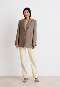 Brun, overdimensioneret blazer i uldblanding med store lommer, enknappet design og guldknapper, parret med cremefarvede bukser og sorte hæle.