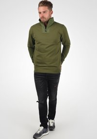 Olivgrüner Quarter-Zip-Pullover aus weichem Stoff, mit geripptem Saum und Seitennähten. Kombiniert mit schwarzen, abgetragenen Jeans und Sneakern.