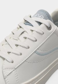 Λευκό δερμάτινο αθλητικό παπούτσι με λευκά κορδόνια, ανοιχτό μπλε ζιγκ-ζαγκ ραφή στο πλάι και γκρι επενδεδυμένη γλώσσα με λευκό λογότυπο κειμένου.