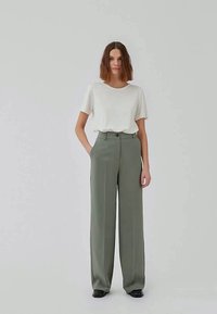 Femme debout portant un haut blanc à manches courtes rentré dans un pantalon large taille haute gris-vert et des chaussures noires, sur fond uni.