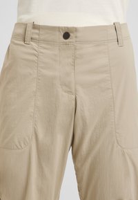 Pantaloni leggeri beige con una texture liscia, dotati di chiusura frontale con bottone, passanti per cintura e dettagli di cucitura discreti.