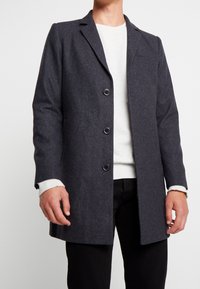 Uomo che indossa un soprabito grigio scuro con bottoni sopra un maglione bianco e pantaloni neri su uno sfondo bianco semplice.