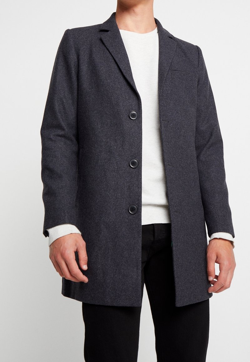 Uomo che indossa un soprabito grigio scuro con bottoni sopra un maglione bianco e pantaloni neri su uno sfondo bianco semplice.