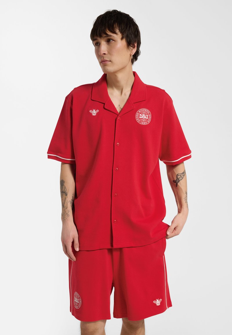 Młoda osoba ubrana w czerwoną koszulę na guziki i krótkie spodenki z białymi logotypami, stojąca na tle jednolitej białej ściany, patrząca na bok.