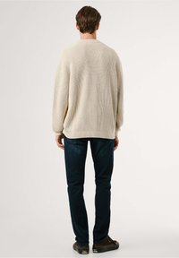 Pull en tricot beige avec un motif texturé, poignets et ourlet côtelés, associé à un jean bleu foncé et des chaussures marron, vu de dos.