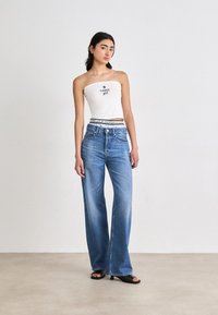 Fehér pánt nélküli felső felirattal, világoskék, bő szárú farmernadrággal párosítva. Simaságot sugalló denim textúra, közép magas derék és pántos dizájnnal ellátott fekete papucsok.