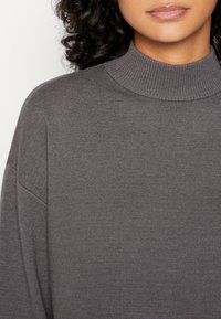 Pull-over gris, avec un col haut à côtes, une texture en tricot doux et des manches droites, soulignant une coupe décontractée.