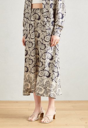 Femme portant une jupe midi beige et marine à motif cachemire avec une veste assortie, un haut blanc et des sandales à talons nude à brides sur un sol en bois.