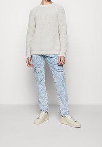 Suéter de punto beige claro con tejido texturizado, combinado con vaqueros desgastados de color azul claro y zapatillas deportivas beige. Ajuste casual y relajado en todo.