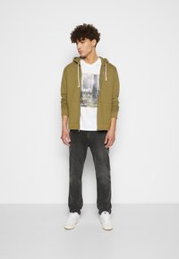 Olivengrønn hettejakke med glidelås og snorer, hvit grafisk T-skjorte, svarte jeans og hvite sneakers med gummisåler. Enkelt design, avslappet passform.