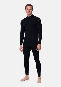 Un uomo è in piedi a piedi nudi indossando un fitted top a maniche lunghe nero e leggings sportivi contro uno sfondo bianco semplice.