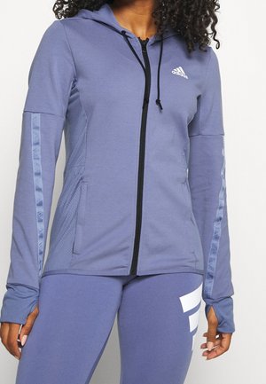 Vrouw draagt een lavendelgekleurde Adidas-jas met rits en kap, duimgaten en bijpassende legging met een wit logo op de linker dij.