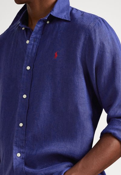 Μπλε λινό πουκάμισο με γιακά που κουμπώνει, άσπρα κουμπιά και μικρό κόκκινο κεντητό λογότυπο στο στήθος. Μαλακή υφή με άνετη εφαρμογή.