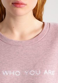 Ljusrosa, kortärmad t-shirt i bomull med rund hals; har vit broderad text: "WHO YOU ARE" på framsidan.
