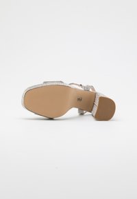 Silberne, Riemchen-High Heelsandale mit einer strukturierten, glitzernden Oberfläche. Verfügt über eine robuste Sohle und einen Knöchelriemen, der ein schlankes Design und eine elegante Form zeigt.