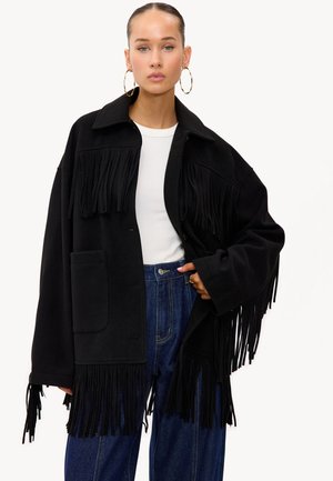 Vrouw in een zwarte franjejasje, witte top, wijde blauwe spijkerbroek en grote gouden oorbellen, posando met één hand in haar zak.