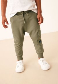 Maslinasto zelene trenirka hlače s rebrastim manšetama i podesivim vezicama, s bočnim džepom za teret i crnom naljepnicom. Nošene s bijelim tenisicama.