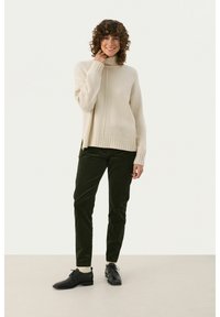 Beige Rollkragenpullover mit geripptem Saum und seitlichen Schlitzen, kombiniert mit dunkelgrünen Cordhosen und schwarzen Schnürschuhen.