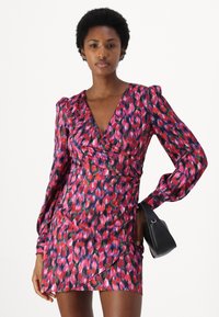 Rochie cu un model roșu și roz, cu decolteu adânc în V, mâneci lungi cu volane și talie ajustată. Acoperită de o geantă mică neagră.