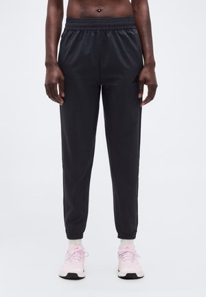 Personne portant un pantalon de jogging noir et des chaussures de course roses avec des chaussettes blanches, debout devant un fond blanc.
