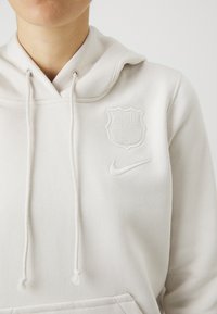 Hoodie gris clair avec cordon de serrage et poche avant. Présente un logo FC Barcelona en relief et un swoosh Nike de couleur assortie.