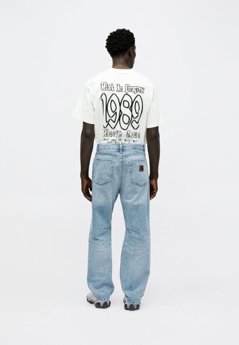 Man wearing white "Work In Progress 1989 World Tour" T-shirt and light blue jeans, standing facing away against plain white background.