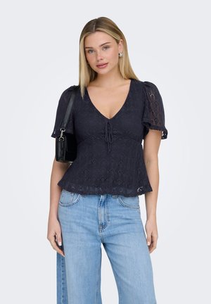 Vrouw draagt een zwart kanten top met korte mouwen en een V-hals, gecombineerd met een blauwe high-waisted spijkerbroek, en houdt een kleine zwarte schoudertas vast.