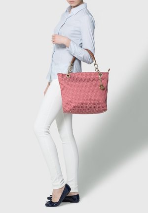 Kvinna i ljusblå skjorta, vita byxor och svarta lägenheter som håller en stor rosa mönstrad axelväska med bruna handtag och ett guldcharm.