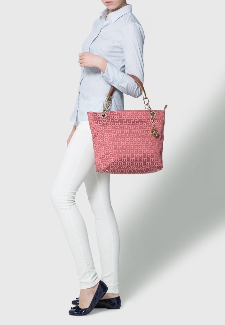 Femme portant une chemise bleu clair, un pantalon blanc et des ballerines noires, tenant un grand sac fourre-tout rose à motifs avec des poignées marron et un charme doré.