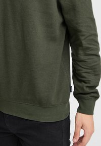 Persona che indossa un maglione a maniche lunghe di colore verde oliva con texture e pantaloni neri, con un piccolo etichetta visibile sulla cucitura laterale del maglione.