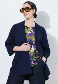 Námořnický cardigan s otevřeným frontem přes vzorovaný zeleno-fialový top. Velké sluneční brýle se žlutým nádechem a zlaté kruhové náušnice. Tmavé džíny.