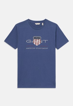 Zilā kokvilnas T-kreklā ar apaļu kakla izgriezumu, kurā ir drukāts logo un vairoga dizains sarkanā un baltā krāsā ar tekstu "1949" un "AMERICAN SPORTSWEAR".