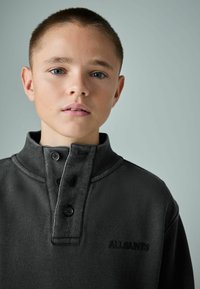 Tamsiai pilkas džemperis su aukštu apykakle, trimis sagomis ir subtiliu siuvinėjimu. Minkšta tekstūra su laisvalaikio stiliumi.