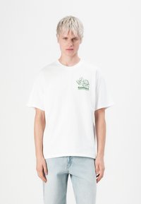 Blonde man in een wit T-shirt met een groene cartoon schildpad en tekst van een elektronicawinkel, gecombineerd met lichtblauwe spijkerbroeken, tegen een witte achtergrond.