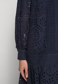 Robe bleu marine avec broderies florales délicates et motifs découpés. Manches longues avec des poignets texturés et un design de jupe à volants.